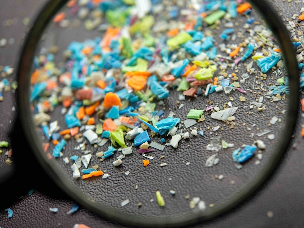 A large black magnifying glass is held over a collection of small colourful plastic particles on a grey surface.