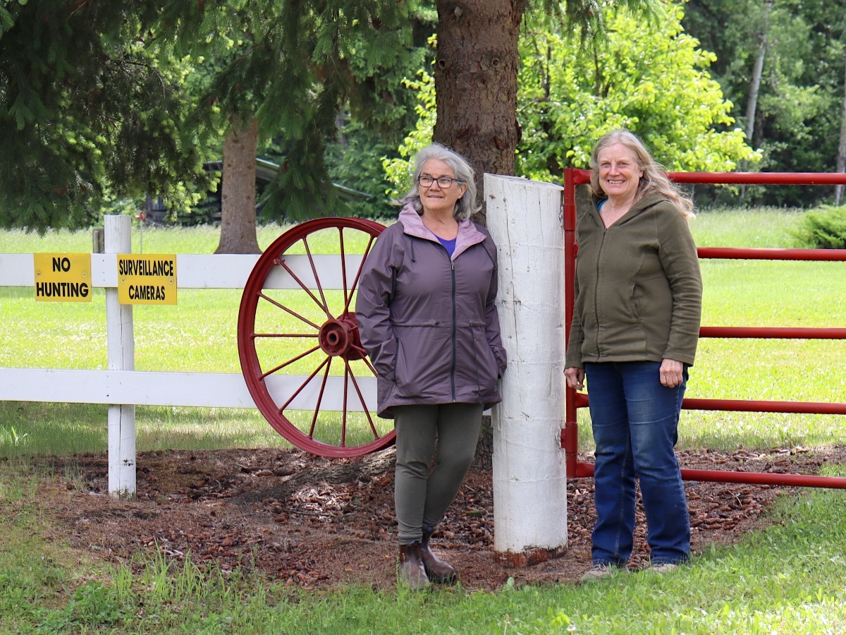 Two women stand next to a red gate and a white fence. A sign declares the presence of surveillance cameras.