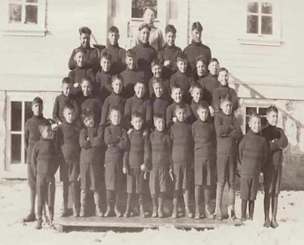 An old black and white photo of a group of schoolboys in black uniforms posing in front of a white building.