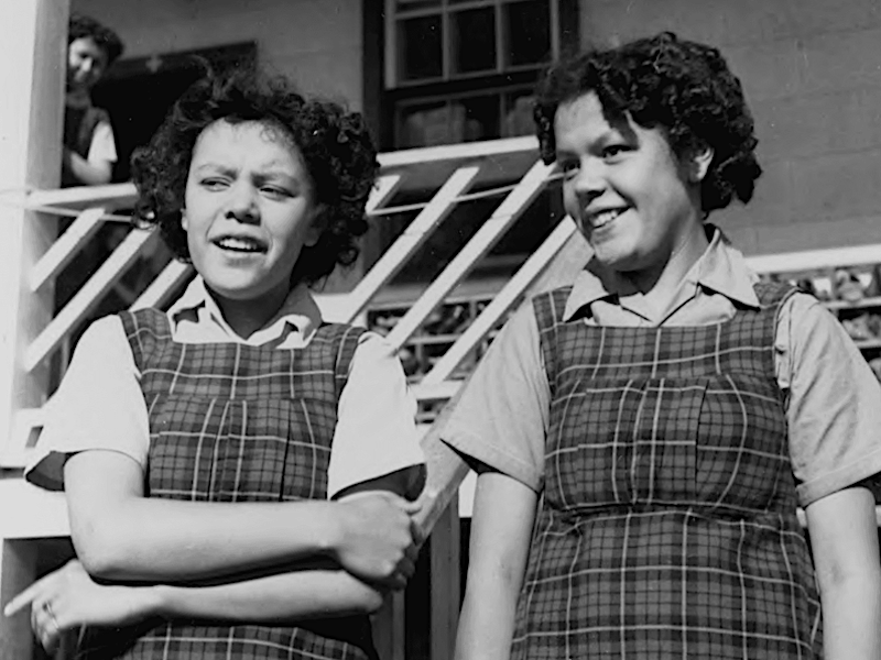 A black and white photo of two schoolgirls in identical uniforms, both with curly dark hair.
