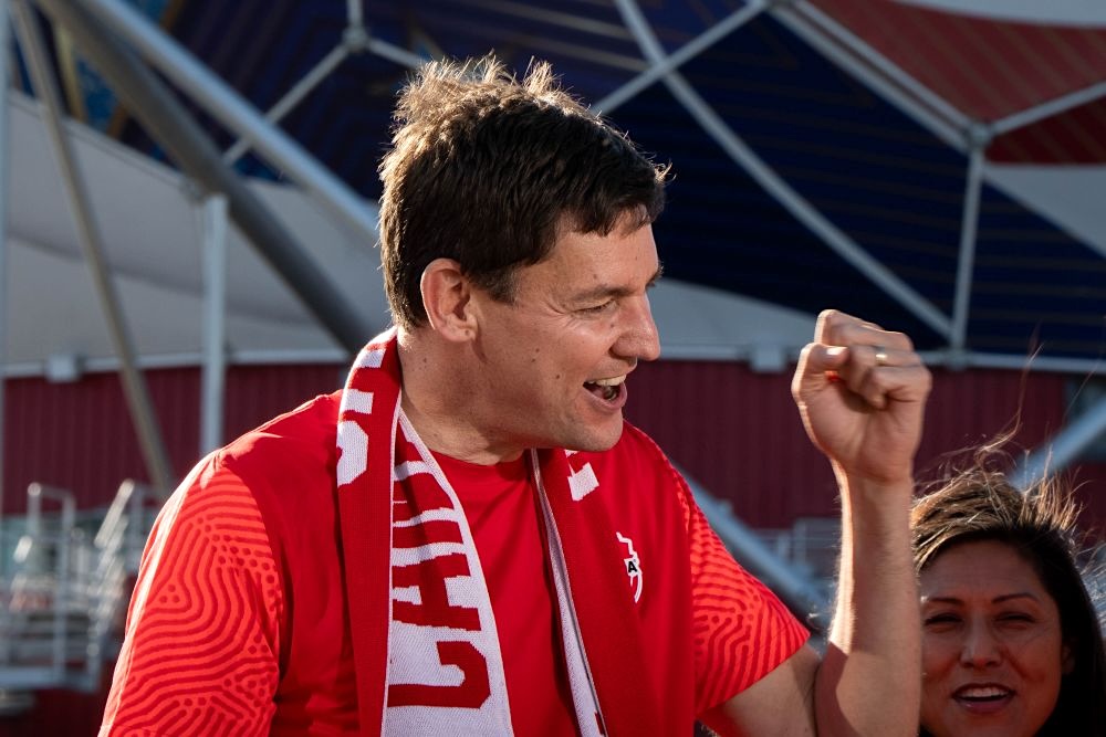 A tall man with light skin tone and short brown hair wears a red soccer jersey and pumps his fist in the air.