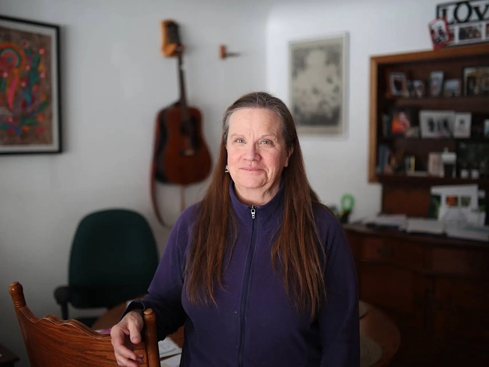 A woman with light skin tone and long straight reddish and grey hair wears a purple zip-up fleece and poses in a room in a home.