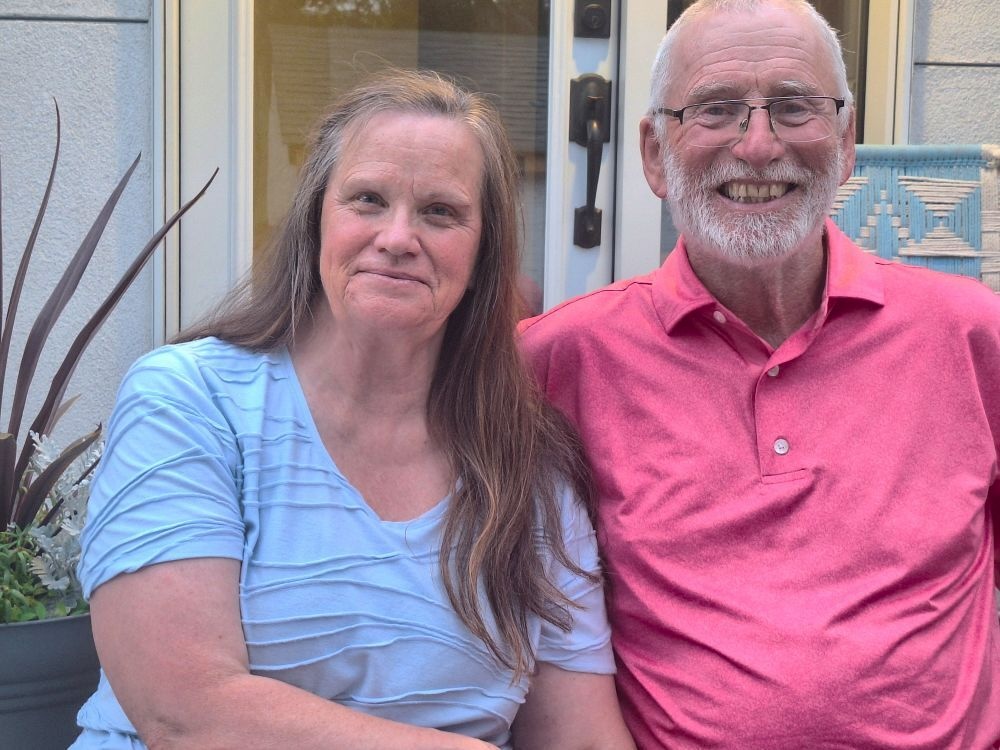 A couple poses for the photo smiling. On the left is a woman with light skin tone and long straight reddish grey hair. On the right is a man with light skin tone, thinning white hair and a white beard.