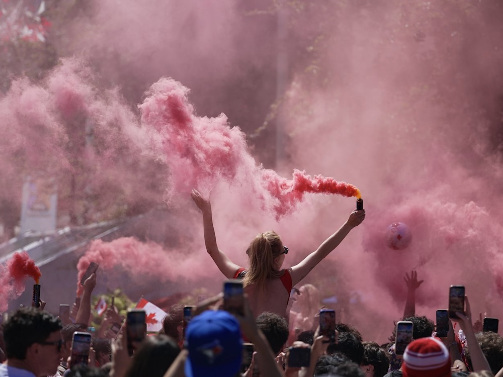 A woman sits on someone&rsquo;s shoulders with her back turned to the camera. She is wearing sunglasses and holding a can of festive red smoke, which fills most of the frame. She is cheering in a crowd of soccer fans celebrating the Canadian national team competing in the 2026 FIFA World Cup.