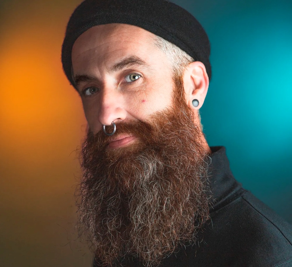 Head shot of a man with light skin tone, a long red beard and a nose ring.