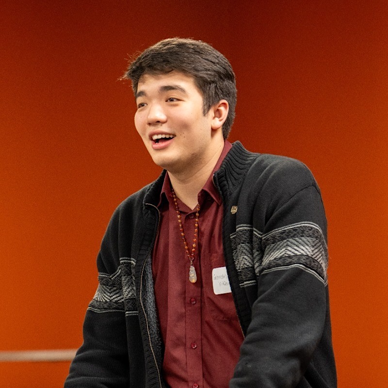 A man in his 20s with light skin and short black hair, wearing a black zip-up cardigan over a dark red collared shirt.