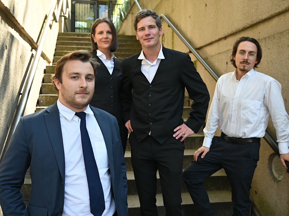 Four people stand in a stairwell. One wears a suit and tie, two wear legal garb and the fourth black pants and a white shirt.