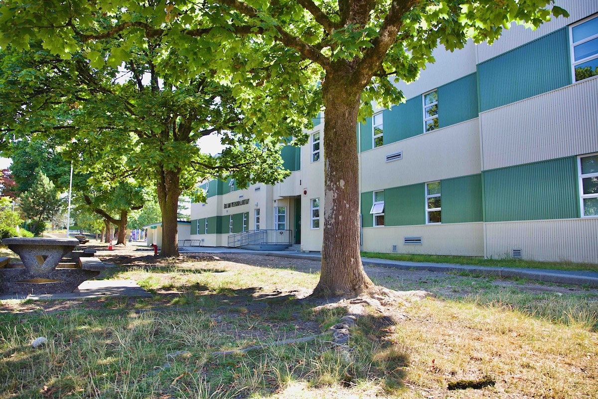 The side view of a multi-storey low-rise building with green and white horizontal siding. Trees line the side of the building and there are concrete tables to the left of the frame.