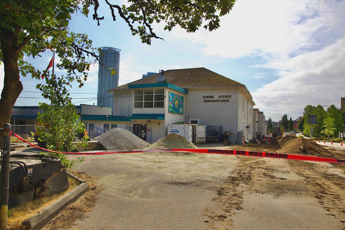 Red caution tape hangs vertically across the frame, blocking a dirt-covered parking lot in front of a two-storey beige school where construction is taking place.