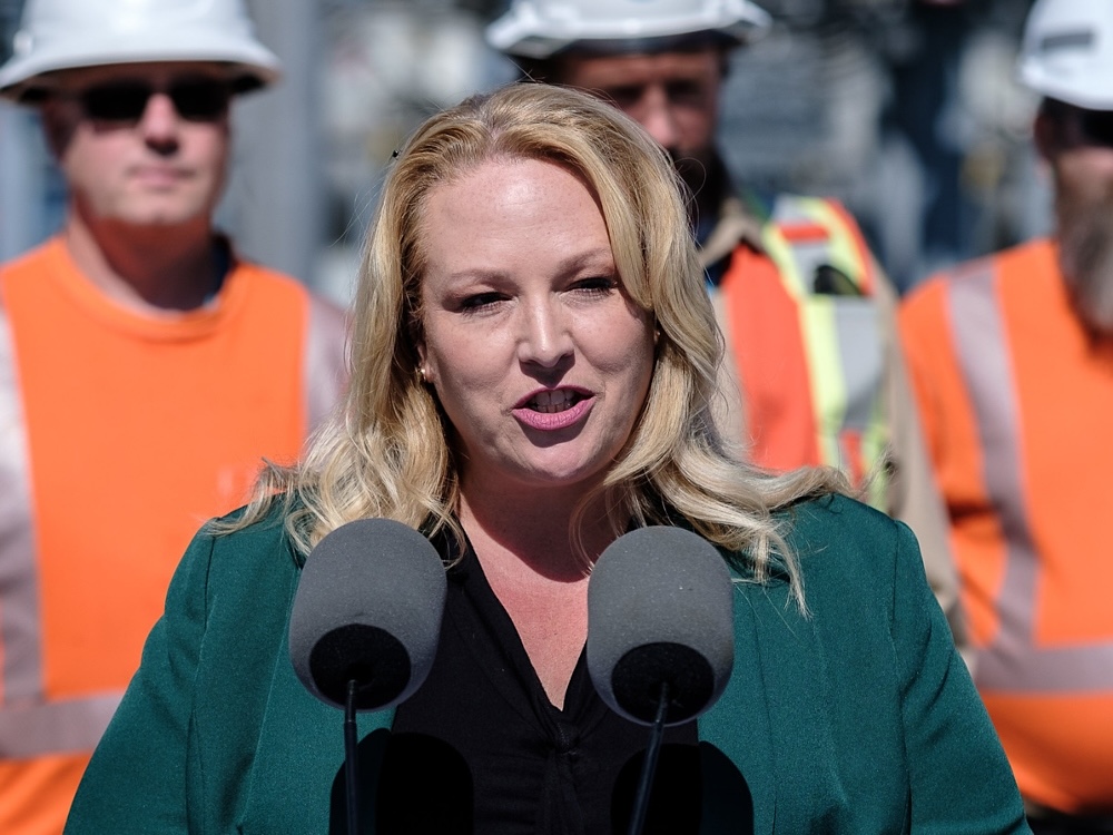 A woman with blond hair and wearing a green blazer speaks into a microphone on a lectern at a press conference.