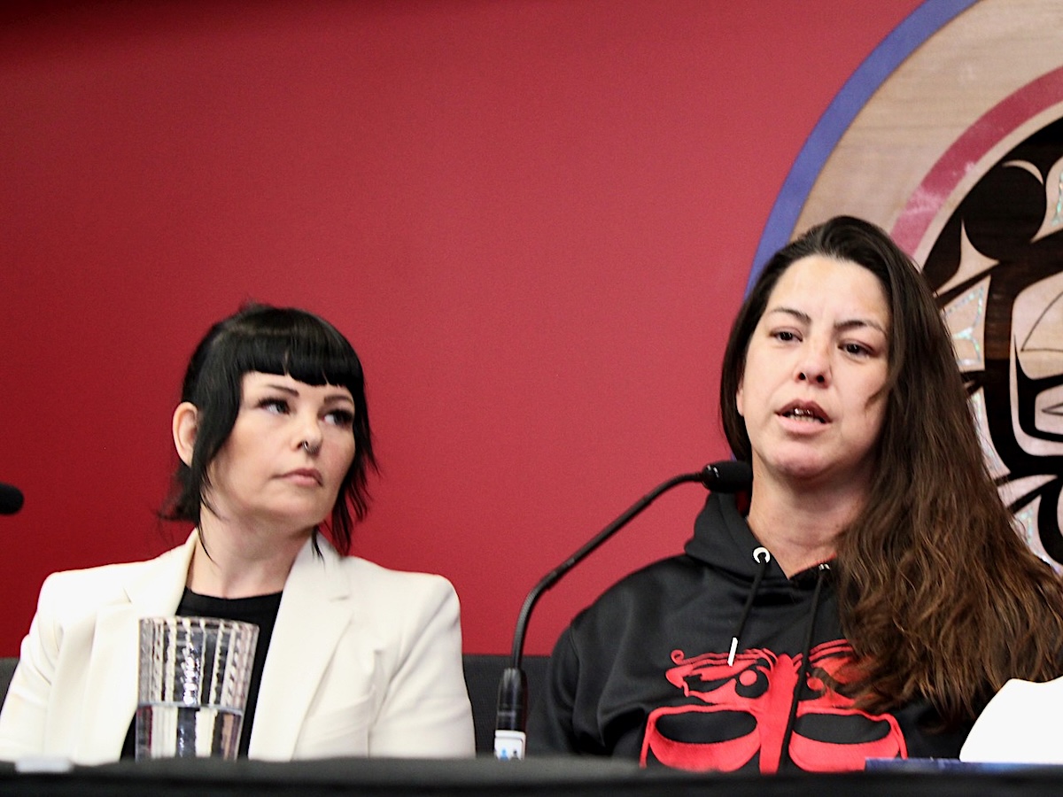 Two women sit at a table with microphones. The woman on the left wears a white suit and has black hair with bangs. The woman on the right has long brown hair and wears a black and red sweatshirt.