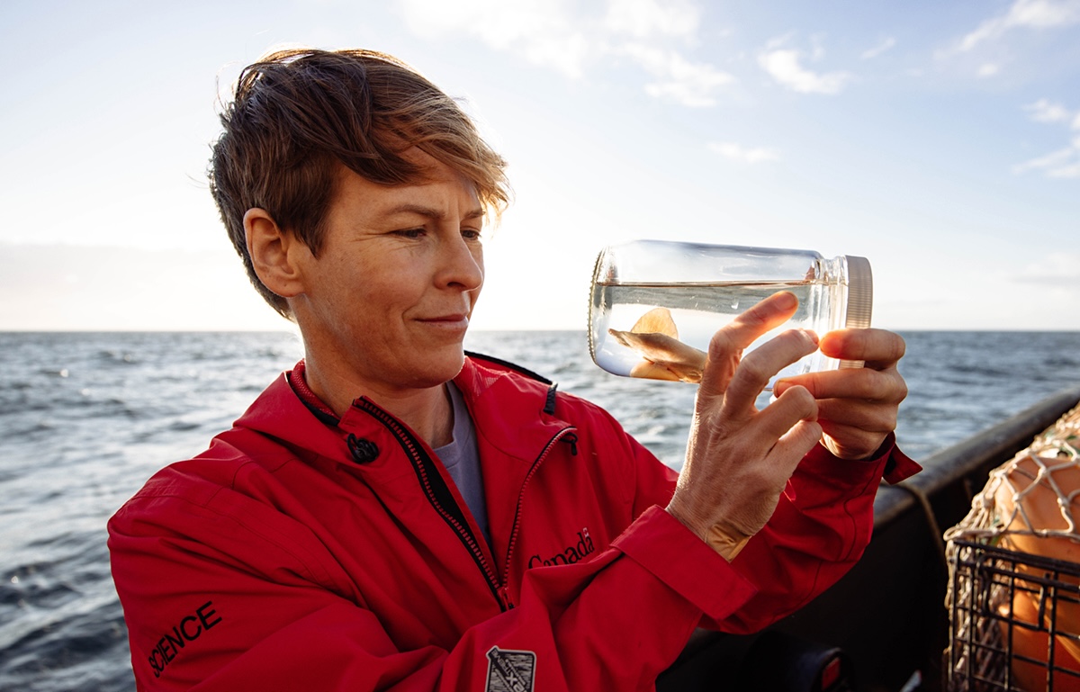 A woman with short blond hair and a red jacket holds up a jar with a small skate specimen in it. She is on a boat and the ocean stretches out to the horizon behind her. 