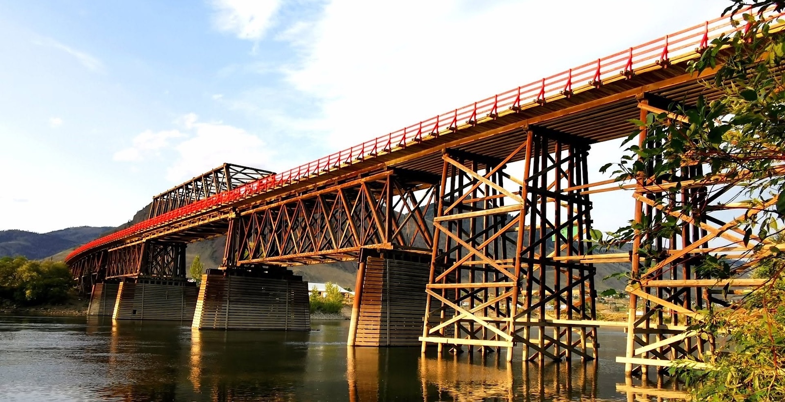 A old wooden bridge with red railing crosses a slow-moving river.
