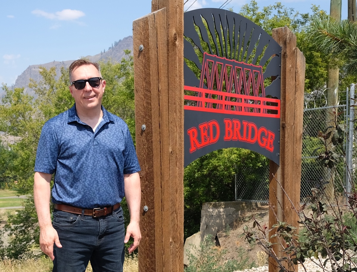 A man with light skin stands beside a metal sign that says &lsquo;RED BRIDGE&rsquo; in red letters. A red bridge is included in the sign, also in red.