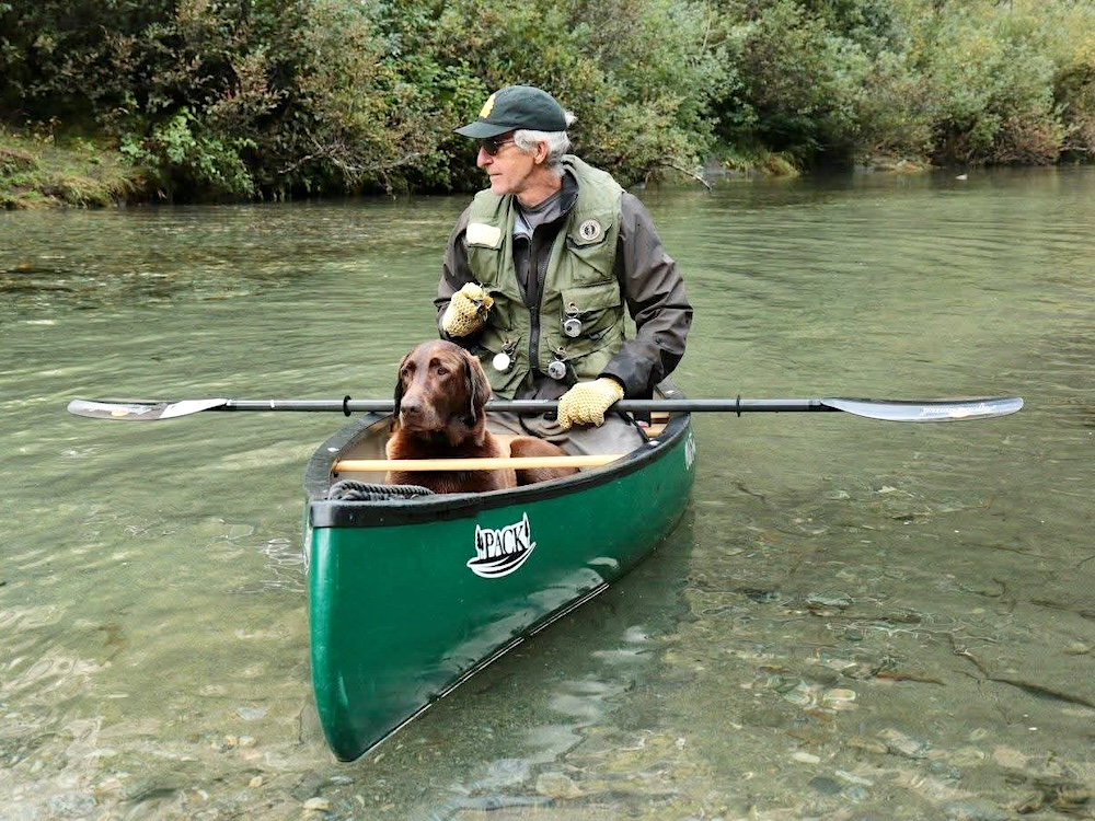 A man in his 70s sits in a canoe, wearing a hat and hiking clothes. A brown Labrador dog sits in the canoe in front of the man.