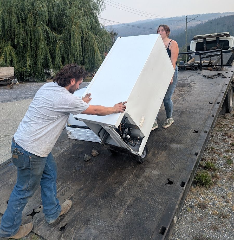 Two people use a dolly to load a used fridge onto a flat-deck trailer.