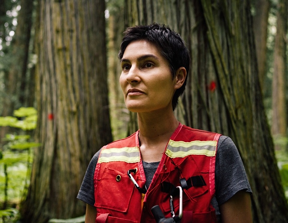A dark-haired woman of early middle age wears a reflective vest and stands in a forest setting.