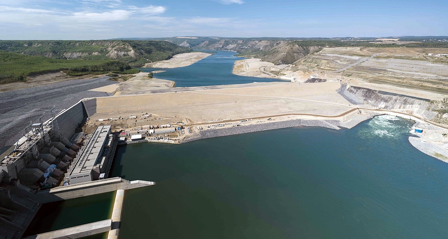 A panoramic photo shows a grey concrete dam with the blue waters of a reservoir behind and river in the foreground.