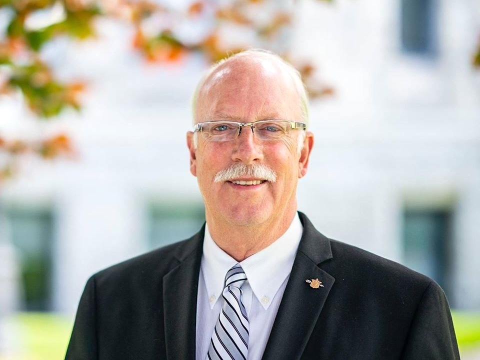 A 60-ish balding man with light-toned skin smiles. He wears glasses, a dark suit and white shirt and tie.