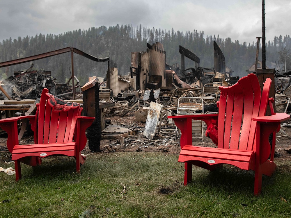Two red plastic Adirondack chairs are partially melted in front of the remains of a burned hotel.