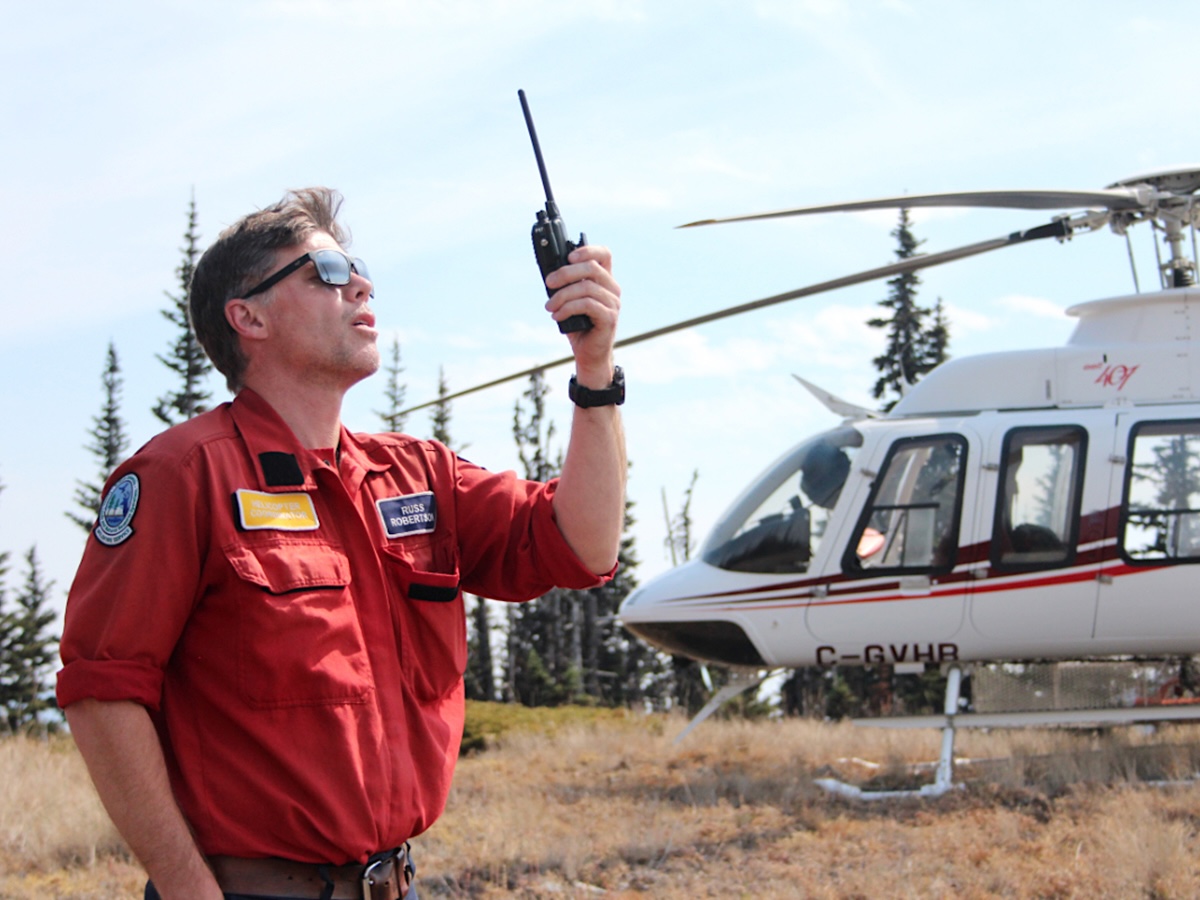A man clad in a red firefighting uniform holds aloft a radio and speaks into it in a grassy meadow. Behind him sits a helicopter.