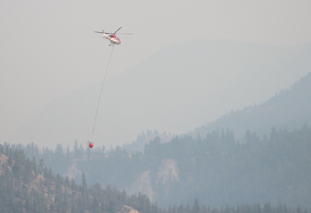 A helicopter carrying a bucket flies through a very smoky valley.