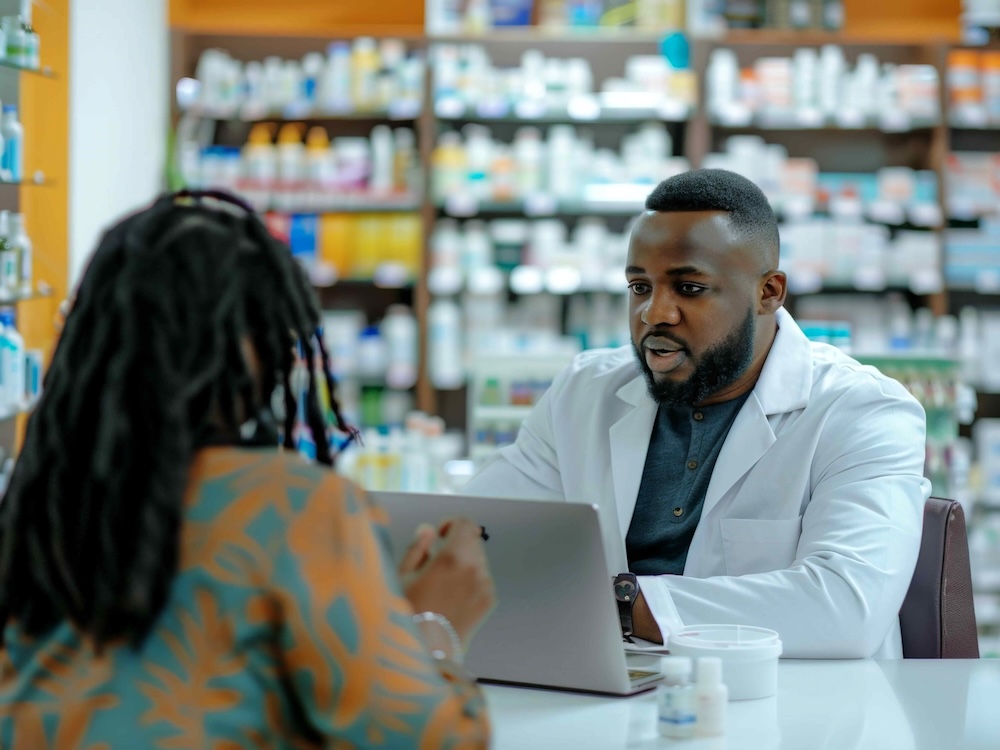 A pharmacist in a white coat speaks to a customer while looking at a laptop, with shelves full of medications in the background.