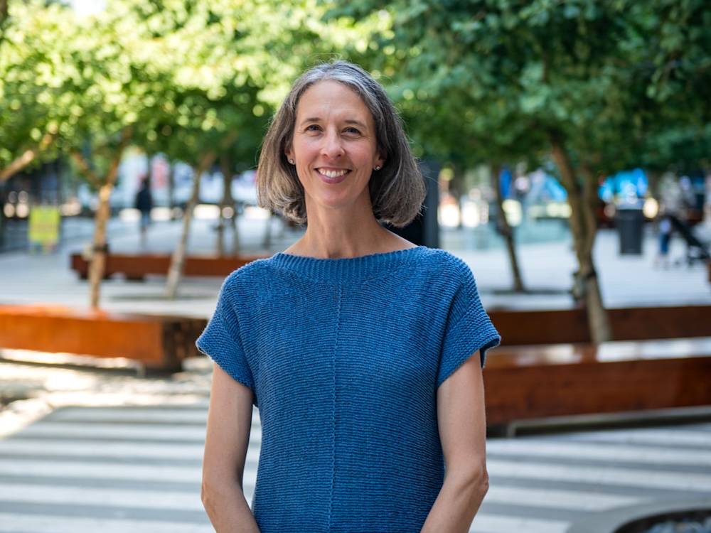 A woman with light skin tone and a greying bob wears a blue short-sleeved sweater.
