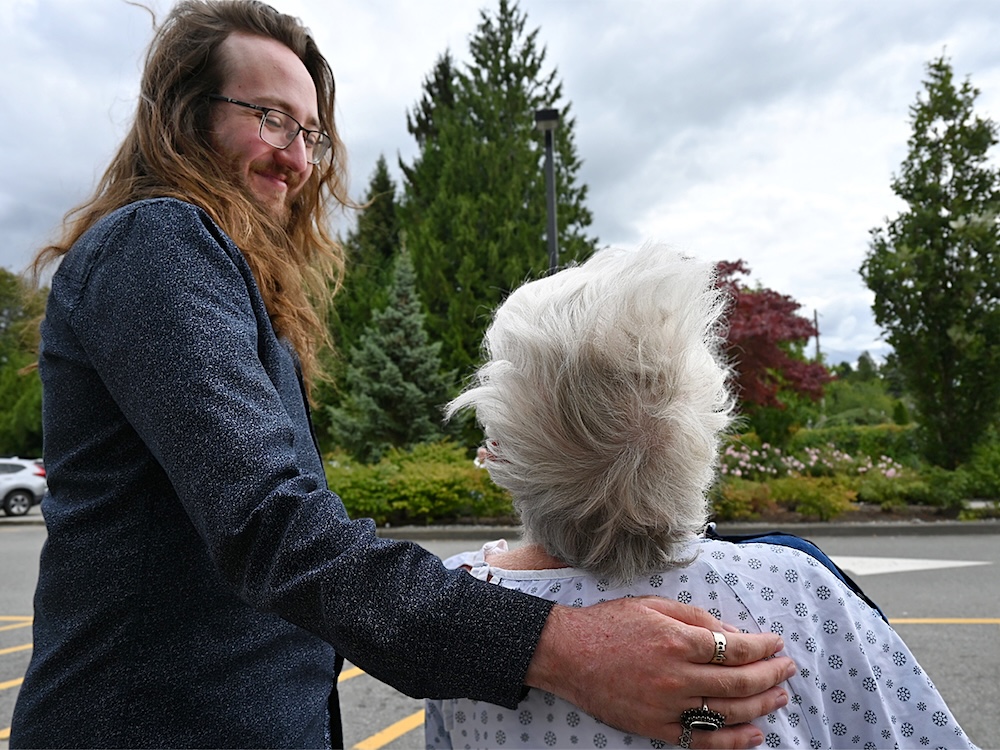 Nathan Davis puts his arm around his grandmother, Elizabeth Davis, who is wearing a hospital gown and seated in a wheelchair.