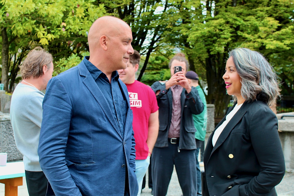A bald middle-aged man gives a sidelong glance while speaking to a middle-aged woman with shoulder-length wavy hair and bright pink lipstick.