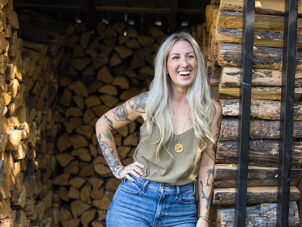 A 40-something woman with long blond hair stands in a firewood storage area smiling.