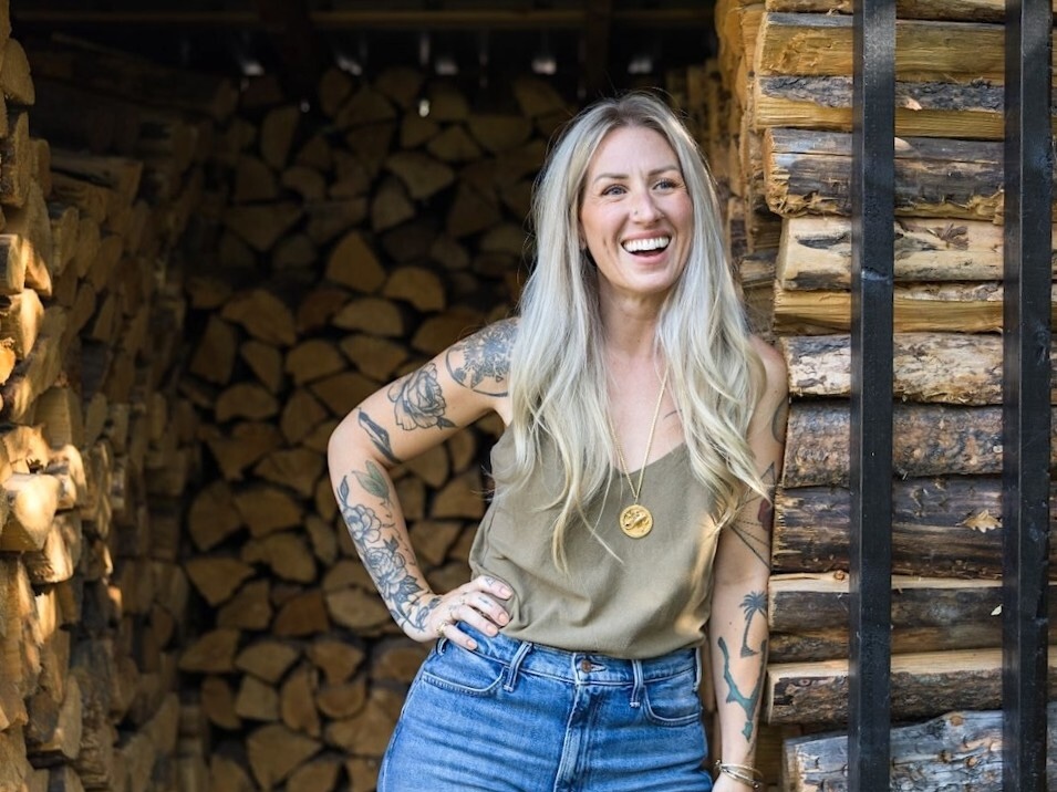 A 40-something woman with long blond hair stands in a firewood storage area smiling.