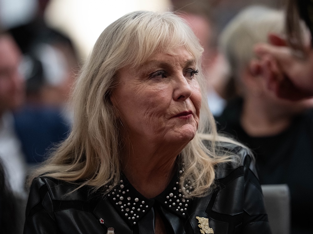 A woman in her 70s with blond hair and bangs looks to the right while seated at an event.