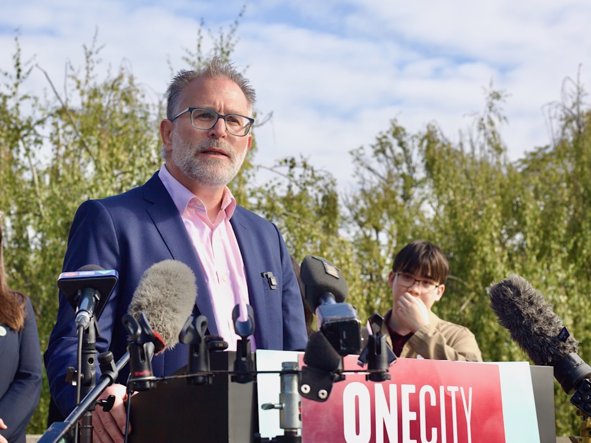 A middle-aged man with short grey hair stands at a lectern that reads &lsquo;OneCity&rsquo; in an outdoor setting.