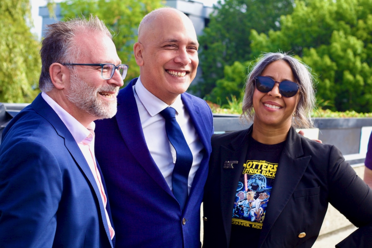 A man with short grey hair and glasses, a bald man and a woman with wavy grey and black hair stand with their arms around one another&rsquo;s shoulders in an outdoor setting. 