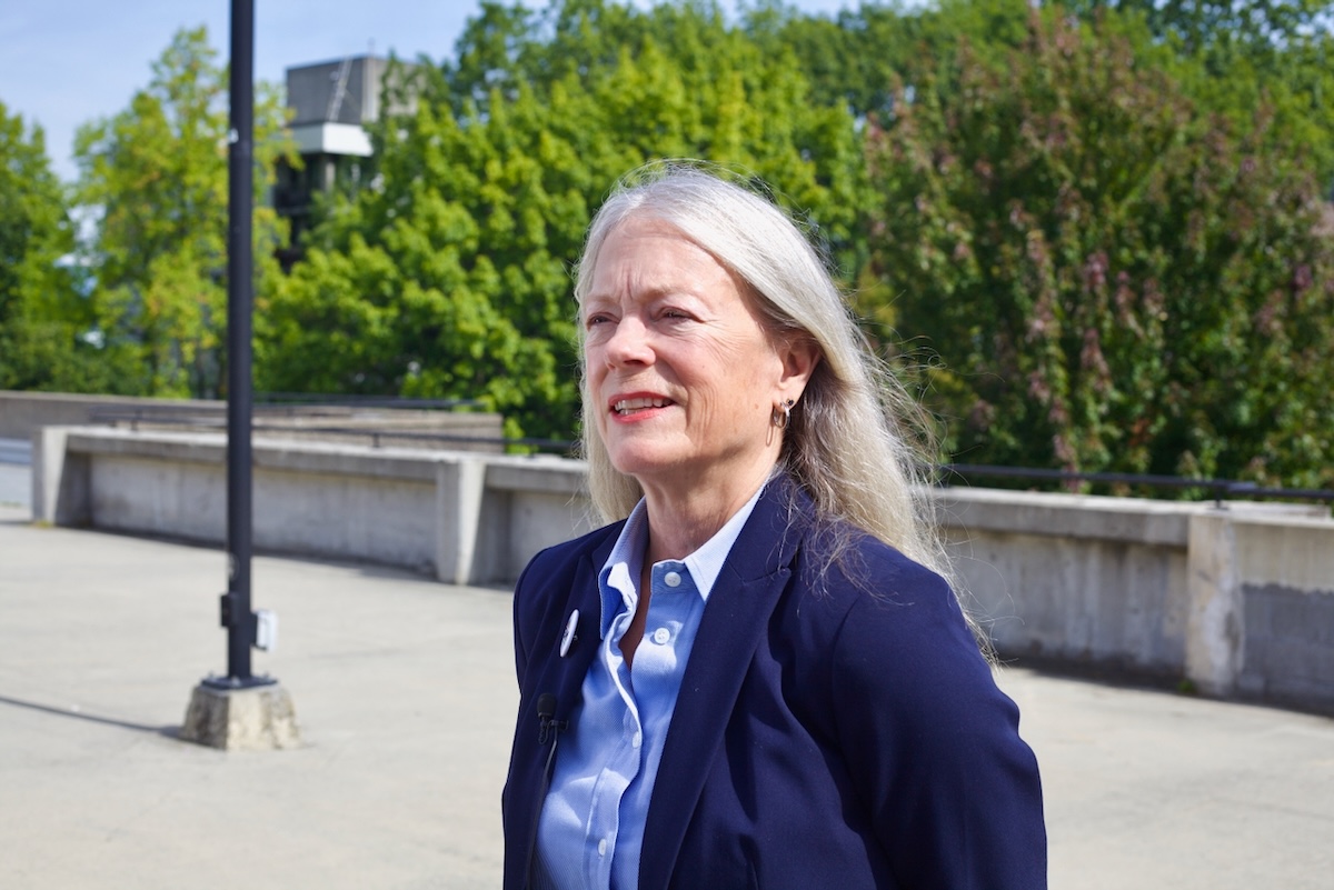 A woman in her 60s with long grey hair stands on a sunny concrete outdoor plaza.