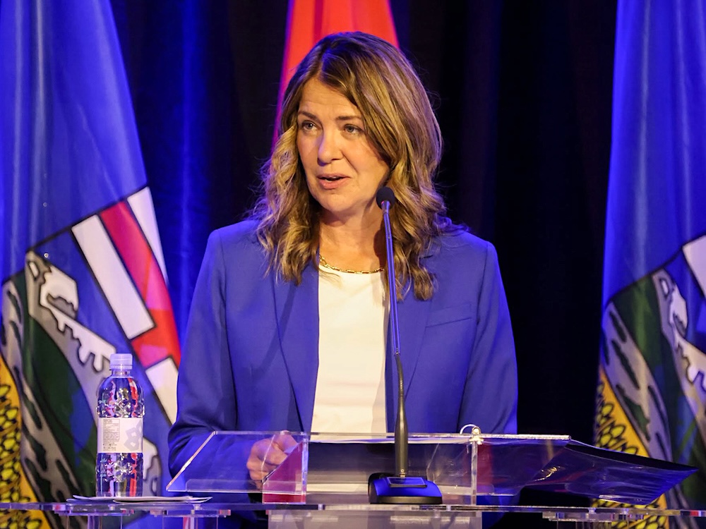 Danielle Smith stands at a lectern wearing a blue blazer. Behind her are two Alberta flags flanking a Canadian flag.