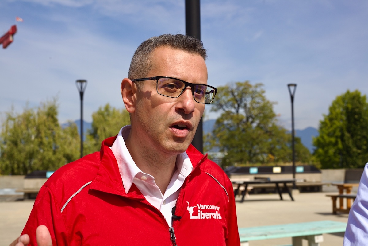 A middle-aged man with curly grey hair and glasses wearing a red athletic zip-up jacket in an outdoor setting.