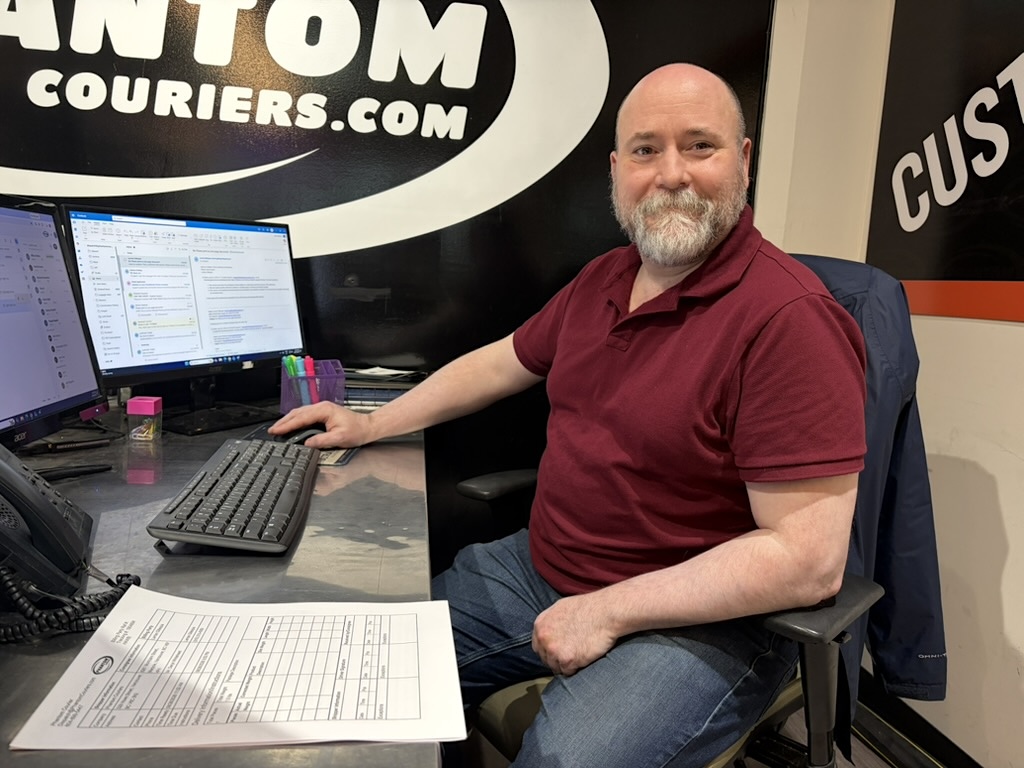 A man in his 50s wearing a red shirt with a grey beard sits at a desk.