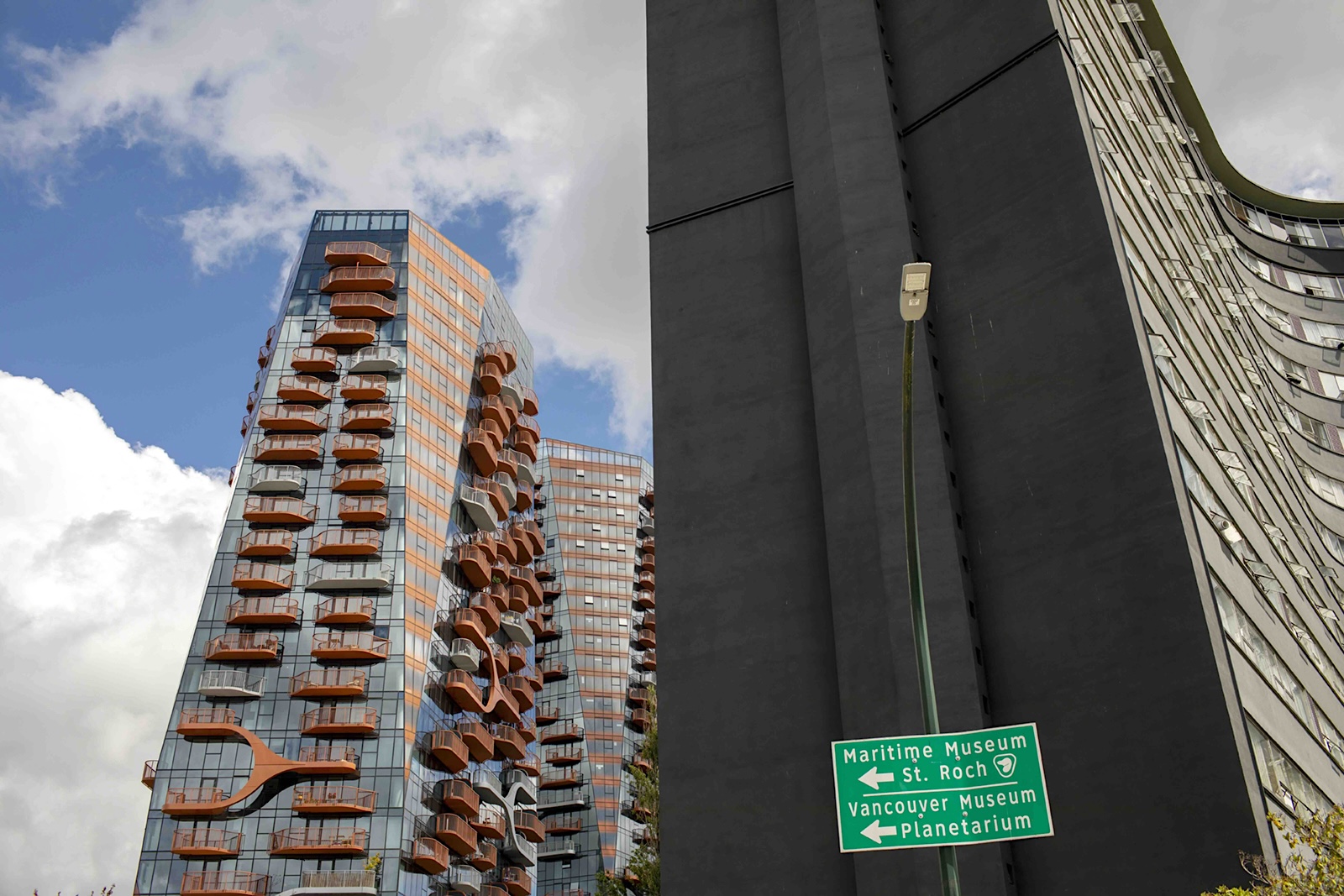 On the left, a view of a contemporary highrise apartment building of the Sen̓&aacute;ḵw development. In the middle of the frame, a green sign that points to the Maritime Museum and Vancouver Museum and Planetarium stands in front of the black side wall of an older apartment building of a similar height.