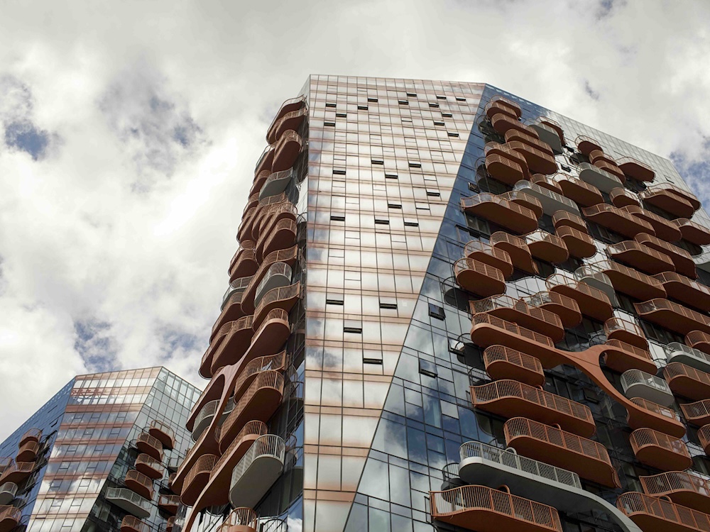Shot from below, a photograph depicts two towers of the Sen̓&aacute;ḵw development; they have bronze accents and balconies. A blue sky with fluffy white clouds is reflected in the glass walls of the buildings.