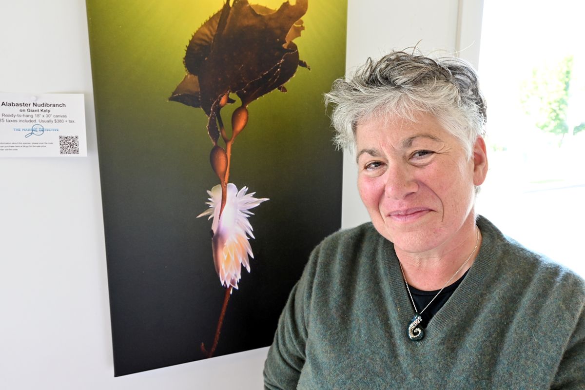 A woman with light skin, short grey hair and a green sweater smiles as she leans against a picture of a pale white nudibranch clinging to a piece of brown kelp.