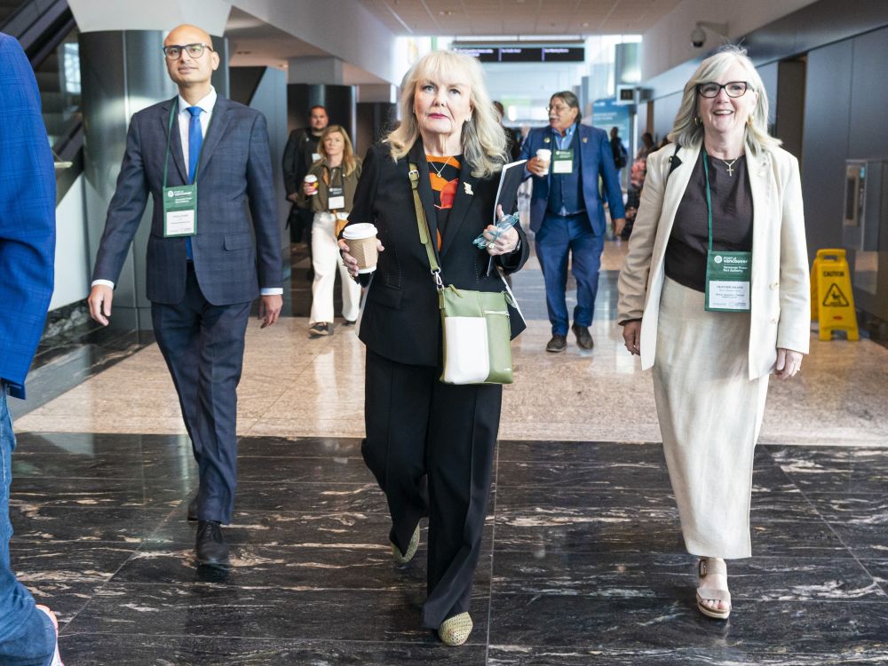 A woman with grey hair and wearing a blazer and dress pants, walks down a hallway flanked by a man and a woman. She&rsquo;s holding a coffee cup.