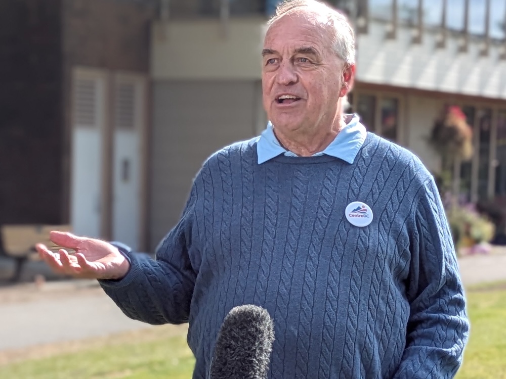 A 60-ish light-skinned man in a blue sweater with a &lsquo;CentreBC&rsquo; pin gestures as he speaks outdoors.