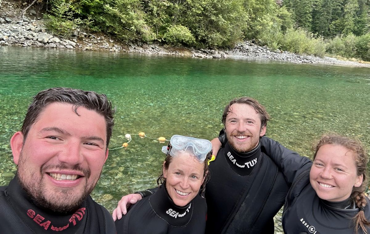 Four people wearing black dry suits grin for the camera while standing on the edge of a green river surrounded by lush green trees.