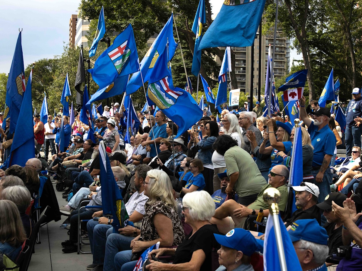 A large crowd of people in blue shirts, many with Alberta flags, outdoors on a plaza.