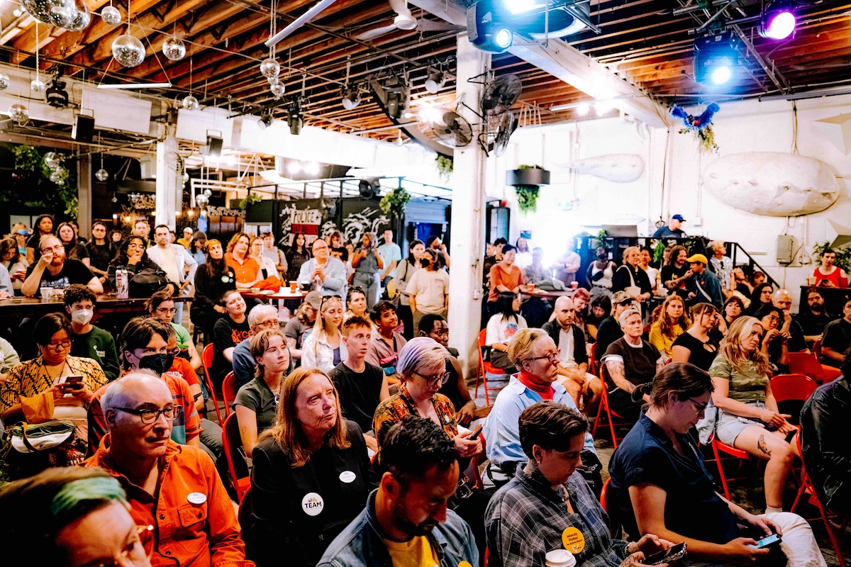 A large audience sits on folding plastic chairs in a brightly lit indoor venue.