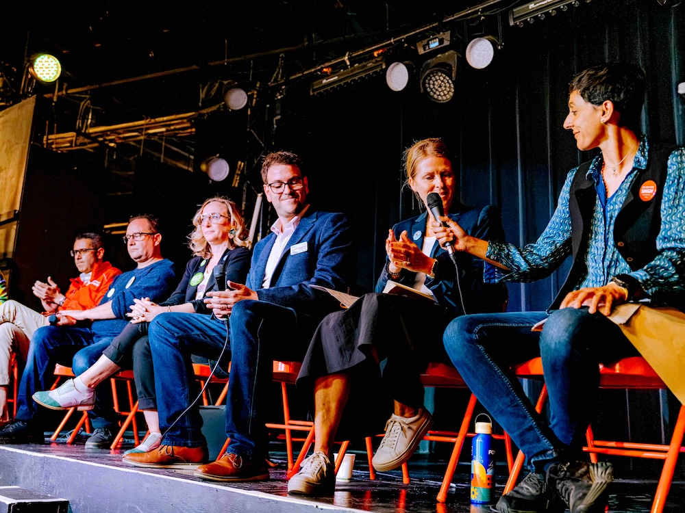 Seven people are seated in a row onstage on red folding chairs. At right, Devyani Singh, with short dark hair and a blue patterned shirt and black vest, passes a microphone to Rebecca Bligh, with long blond hair tied back and a denim jacket over a white shirt.