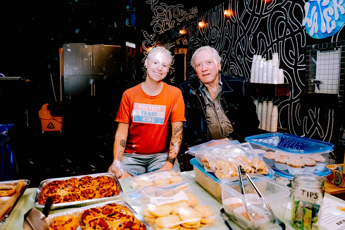 Two people with light hair sit behind a table loaded with baked goods. 