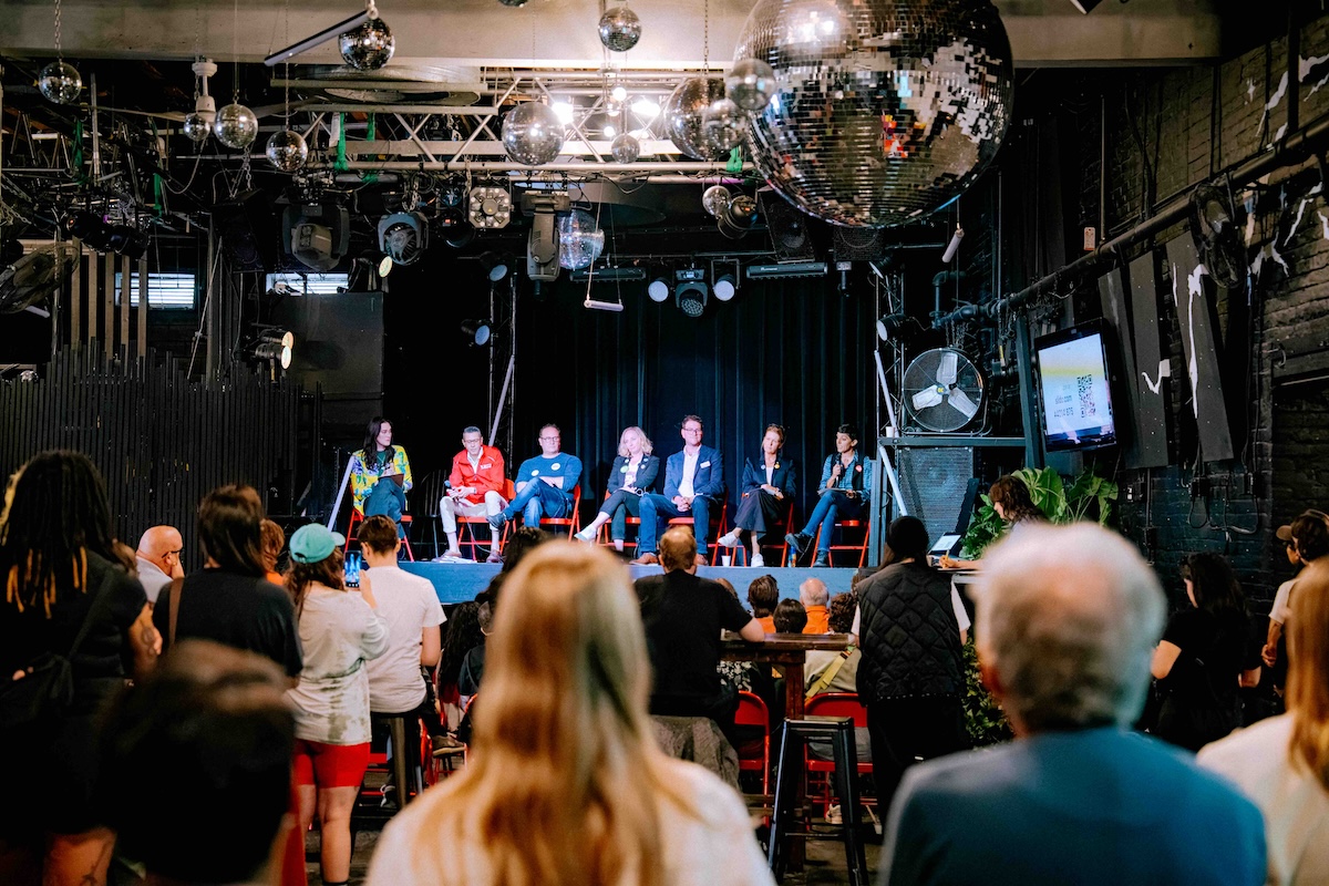 Disco balls hang from the ceiling of an event venue where an audience, shot from the back, watches a panel of municipal candidates seated on red folding chairs onstage.